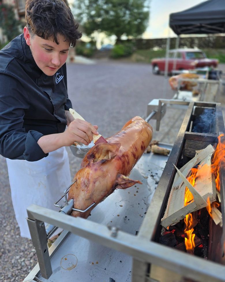 Jeune cuisinier en veste Chedru qui badigeonne un cochon de lait embroché au-dessus des braises
